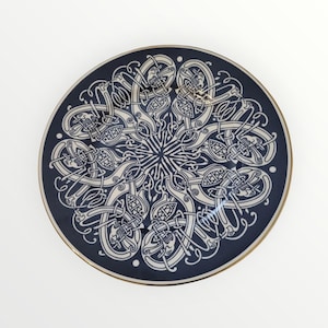 May include: A round, navy blue decorative plate with a gold rim. The plate features a detailed, symmetrical white Celtic knot design in the center and around the edges. The design is intricate and appears to be hand-painted.