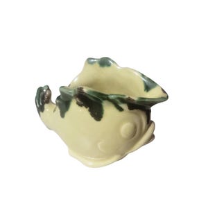 May include: A small ceramic creamer shaped like a fish with a cream-colored body and dark green accents along the top rim and tail. The vessel features a wide, open mouth that serves as the pouring spout and a molded eye on the side.