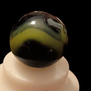 May include: A spherical glass marble featuring a deep black base color with a prominent, swirling band of opaque lime green glass running horizontally across the center of the surface, creating a smooth and reflective finish.