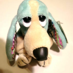 May include: A plush toy dog with large, black eyes and a sad expression. The dog has light blue fur on its head and ears, with a tan snout and paws. The ears have colorful patterned fabric on the inside.
