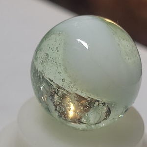May include: A clear glass marble with a white, swirling interior. The marble has a light green tint and is filled with tiny bubbles. The bottom of the marble has a reflective, metallic appearance.