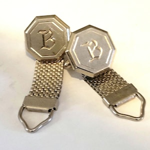 May include: A pair of silver-toned cufflinks. Each cufflink features an octagonal face with a stylized letter "B" and a mesh strap. The cufflinks have a loop at the end of the strap.