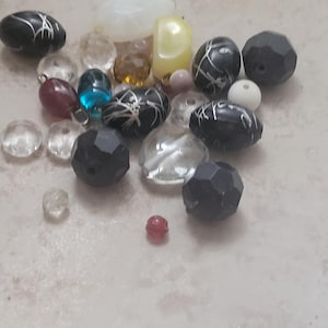 May include: A collection of various beads in different shapes, sizes, and colors. The beads include clear faceted, black with silver designs, and opaque white, yellow, and red beads. Some beads are round, while others are oval or faceted.
