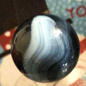 May include: A close-up of a glass marble with a swirling pattern of white and light blue against a dark blue background. The marble is round and glossy, reflecting light.