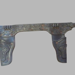May include: A brown leather gun belt with two holsters, both with intricate floral designs. The belt is decorated with a silver-toned buckle.