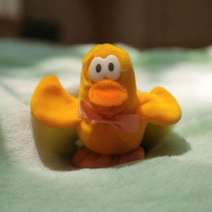 May include: A plush yellow duck toy with a bright orange beak and feet, and large black and white eyes. The duck has outstretched wings and a pink bow tie. The toy is sitting on a soft, light green surface.