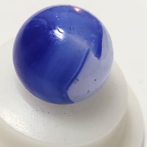 May include: A close-up of a blue and white marble. The glass marble has a swirl pattern of deep blue and white. The marble is resting on a white surface. The marble is a sphere.