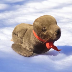 May include: A plush brown seal toy with a red ribbon around its neck, resting on a snowy surface. The seal has a detailed face and a playful, open-mouthed expression. The toy is designed to resemble a real seal.