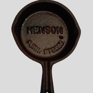 May include: A small black cast iron skillet with a handle. The skillet has the words "Henson  Funiture Stores" embossed on the bottom.
