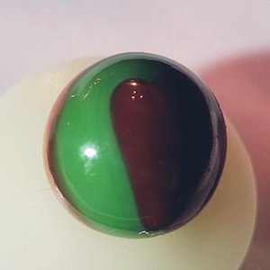 May include: A glass marble with a glossy finish. The marble is divided into two sections: one half is green, and the other is a deep burgundy color. The marble is resting on a white surface.