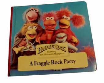 May include: A blue book with a yellow label that says "Fraggle Rock Starring Jim Henson's Muppets" and "A Fraggle Rock Party". The cover features four Fraggles: Red, Yellow, Blue, and Green.