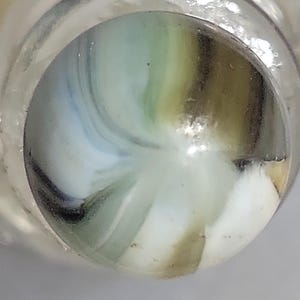 May include: A close-up of a glass marble with a swirling pattern of white, blue, green, and brown colors. The marble is encased in a clear glass container, creating a magnifying effect. The marble's design is reminiscent of a galaxy.