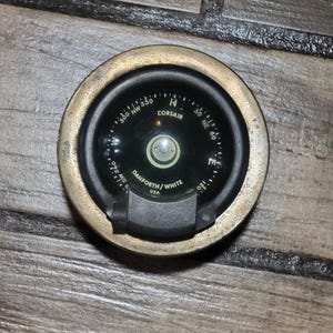May include: A vintage Corsair compass with a brass bezel. The compass face is black with white markings for direction and degrees. The text "CORSAIR" and "DANFORTH/WHITE USA" are visible. A classic navigational tool.