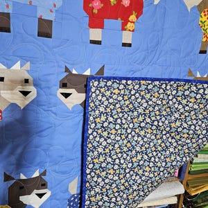 May include: A blue quilt with a patchwork design featuring goats wearing different patterned clothes. The quilt is folded over a stack of fabric scraps in various colors and patterns.