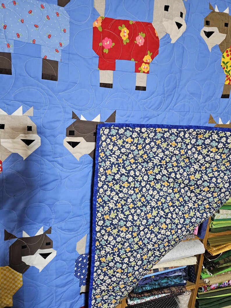 May include: A blue quilt with a patchwork design featuring goats wearing different patterned clothes. The quilt is folded over a stack of fabric scraps in various colors and patterns.