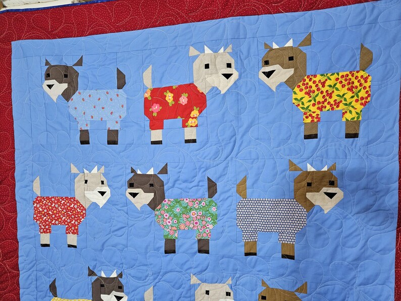 May include: A blue quilt with a red border features a pattern of nine goats wearing pajamas. The goats are made of fabric with different patterns, including floral, polka dot, and geometric designs.