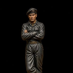 Puede incluir: Figura detallada de una persona con uniforme militar, que lleva una boina con un emblema de cruz. La figura tiene los brazos cruzados, botas y un cinturón. La figura está de pie sobre una superficie texturizada con un fondo negro.