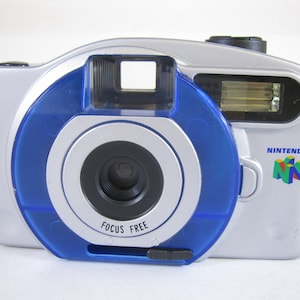 May include: A silver and blue Nintendo 64 camera with a blue lens ring and the words "Focus Free" printed on it. The camera has a built-in flash and a strap.