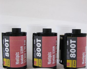 May include: Three rolls of Kodak 800T 35mm motion picture film. The film canisters are black with red and yellow labels. The labels display the film type, brand, and specifications. White plastic film canister lids are in the foreground.
