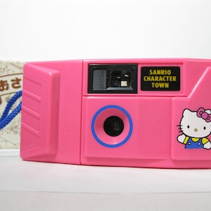 Puede incluir: Una cámara de juguete de plástico rosa con un personaje de Hello Kitty en el lateral. La cámara tiene un visor negro y un anillo azul alrededor del objetivo. El texto "SANRIO CHARACTER TOWN" está impreso en la cámara.