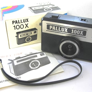 May include: A vintage Pallux 100X camera with its original box and instruction manual. The camera is black and silver with a prominent lens and the words "Pallux 100X" printed on the front. A black wrist strap is attached.