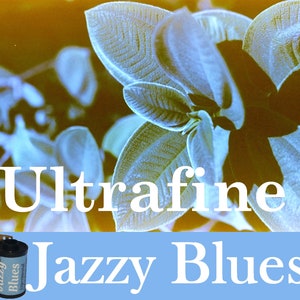 May include: A close-up of a film canister with the text "Jazzy Blues" and "Ultrafine" printed on it. The canister is on a blue background with a blurry image of green leaves in the background.