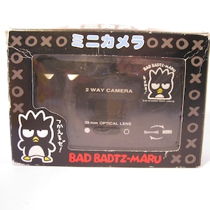 May include: A black and white camera box with a cartoon penguin character on the front. The box says "Bad Badtz-Maru" and "Mini Camera" in Japanese. The camera has a 28 mm optical lens and a panorama/normal setting.
