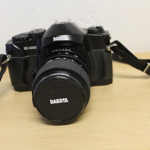 May include: A black Dakota RZ-2000 camera with a lens cap marked "DAKOTA". The camera has a black strap attached. The camera is on a light brown surface.