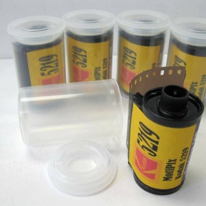 May include: A collection of Kodak MotiPix 5219 film rolls in clear plastic containers. The film rolls have yellow labels with red and black text. One roll is partially unrolled, revealing the film strip.