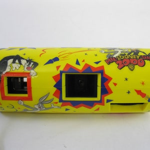 May include: Yellow plastic camera with a black strap. The camera has a cartoon design featuring Bugs Bunny and Sylvester the Cat. The text "MILLOONEY.COM 2000" is printed on the camera.