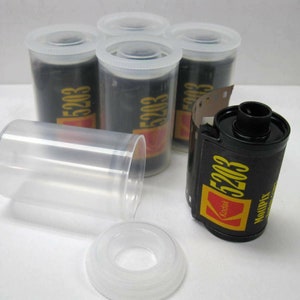 May include: Five rolls of Kodak 5203 black and white film in clear plastic canisters with yellow and black labels. One roll is partially unwrapped, showing the film inside.