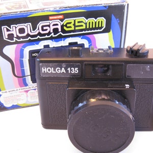May include: A black Holga 135 35mm film camera with a lens cap and a strap. The camera is sitting on a white surface. The camera box is in the background.