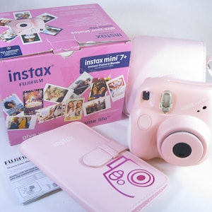 May include: A pink Fujifilm Instax Mini 7+ instant camera with a pink case and a pink photo album. The camera has a lens cover and a flash. The case has a strap and a zipper. The album has a camera icon on the front.