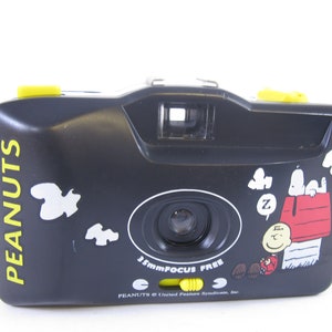 May include: A black 35mm focus free camera with a Peanuts design featuring Snoopy and Charlie Brown. The camera has a yellow strap and yellow buttons.