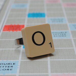 May include: A beige Scrabble tile with the letter "O" and the number "1" printed on it. The tile is on a gray and white game board.