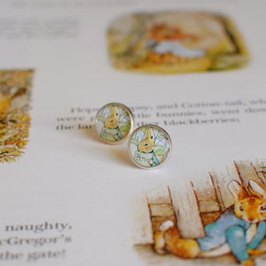 May include: A pair of silver stud earrings featuring a Peter Rabbit illustration. The earrings are round and have a clear glass dome over the image.