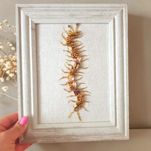 May include: A framed wall art piece featuring a centipede-shaped design crafted from amber, gold, and iridescent beads and crystals. The insect-like figure is centered on a light-colored fabric background within a distressed white wooden frame.