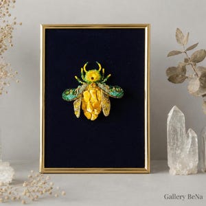 May include: A framed artwork featuring a three-dimensional insect sculpture centred on a dark blue velvet background. The insect is crafted with yellow amber-like stones for the abdomen, green and yellow beadwork for the wings, and a head.