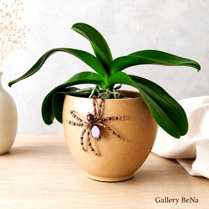May include: A smooth, tan ceramic planter containing a green orchid plant. A decorative spider ornament crafted from purple and gold beads is attached to the front of the pot. The text Gallery BeNa is visible in the bottom right corner.