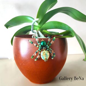 Pode incluir: Um ornamento em forma de aranha feito de contas, com uma conta grande de cabeça verde metálica e um corpo de vidro multicolorido com padrões. As oito pernas são feitas de pequenas contas verdes e cor de pêssego translúcidas. Texto Gallery BeNa.