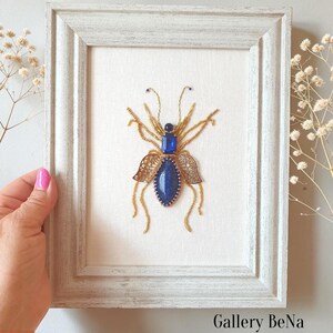 Può includere: Un'opera d'arte da parete incorniciata raffigurante un insetto realizzato con perline. L'insetto presenta un corpo in pietra blu, una testa rettangolare blu e zampe e ali in filo dorato. L'opera è montata su uno sfondo in tessuto bianco.