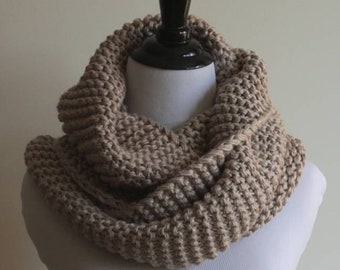 May include: A beige knitted infinity scarf on a white mannequin. The scarf is made with a simple stitch pattern and has a relaxed, drapey look. The text "Pattern" and "By, Winter White Knits" is printed below the scarf.