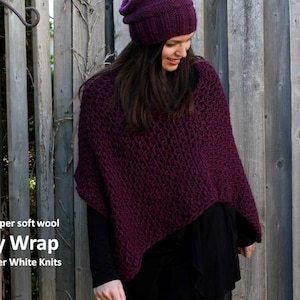 May include: A purple knitted wrap made from 100% super soft wool. The wrap is worn by a person standing in front of a wooden fence. The text "Cozy Wrap" and "by Winter White Knits" is printed below the wrap.