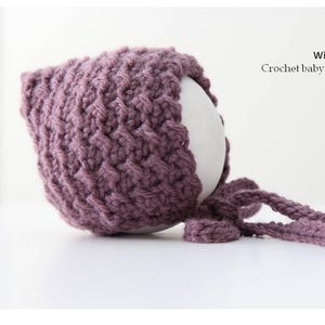 Könnte beinhalten: Gehäkelte Babyhaube in einem gedämpften Lila. Die Haube hat ein strukturiertes Stichmuster und lange Bindebänder. Der Text auf dem Bild lautet "Winter White Knits Crochet baby bonnet pattern."
