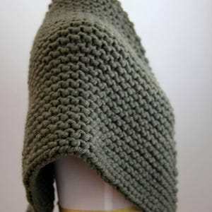 May include: A chunky knit shawl in a solid olive green colour. The garment features a prominent garter stitch texture created with thick yarn, draped over a display form. The design is sleeveless and covers the shoulders and upper torso area.