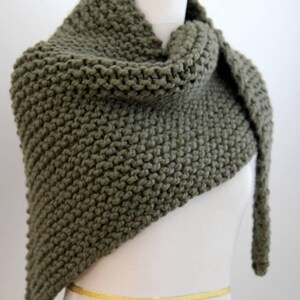Peut inclure: Un châle triangulaire en tricot épais de couleur vert olive uni. L'accessoire présente une texture en point mousse prononcée sur toute sa surface, avec une extrémité drapée plus longue que l'autre, créant une silhouette asymétrique.