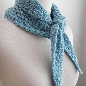 May include: A light blue knitted scarf with a textured, bumpy stitch pattern. The scarf is tied in a simple knot at the front, featuring a triangular shape that drapes around the neck. The yarn has a matte finish and a consistent, solid color.