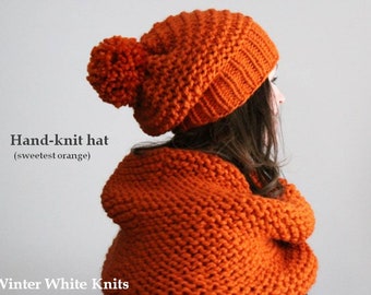 May include: Hand-knitted orange beanie with a pom-pom on top. The beanie is made with a chunky yarn and has a slouchy fit.