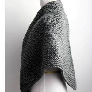 Peut inclure: Un poncho tricoté gris présentant un motif en point mousse épais et texturé. Le vêtement est drapé sur un mannequin blanc, mettant en valeur un col montant et une conception ouverte sur les côtés qui tombe en une silhouette structurée.