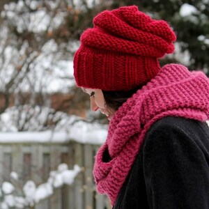 May include: A bright red chunky knit beanie with a ribbed brim and a matching deep pink chunky knit infinity scarf worn over a dark coat. The accessories feature a thick, textured yarn construction designed for cold weather wear.